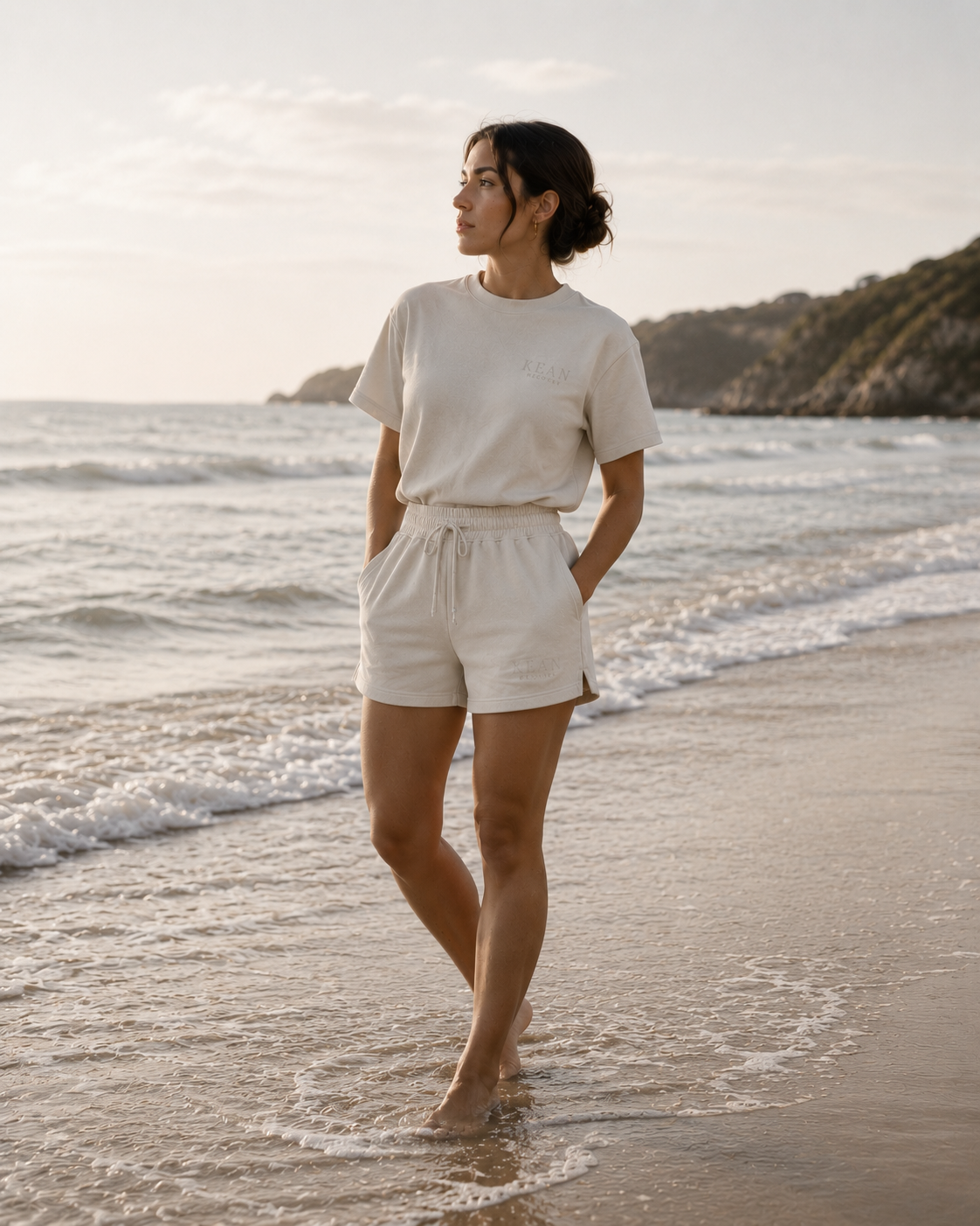 Woman in a white outfit standing on a beach with ocean waves and a mountain in the background. 100% GOTS-certified organic cotton